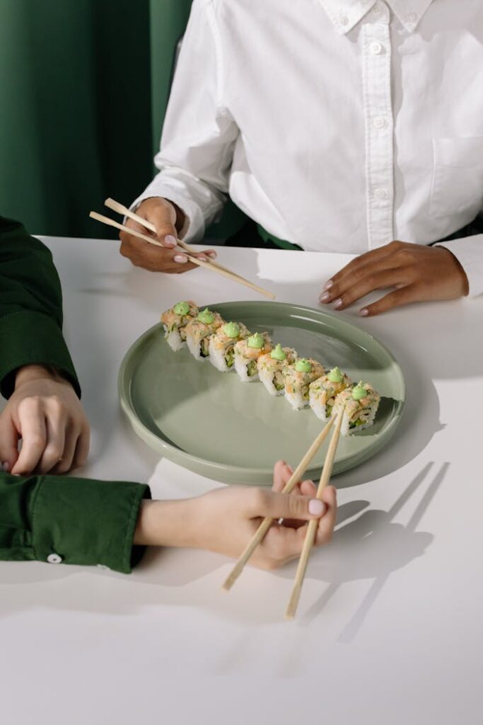 Two individuals using chopsticks to pick sushi rolls from a green plate, creating a vibrant dining scene.